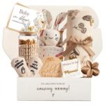 An elegant gift box containing a silver necklace, glass cup, flower hair clip, bunny lovey, baby bath towel, baby socks, and 