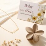 An elegant gift box containing a silver necklace, glass cup, flower hair clip, bunny lovey, baby bath towel, baby socks, and